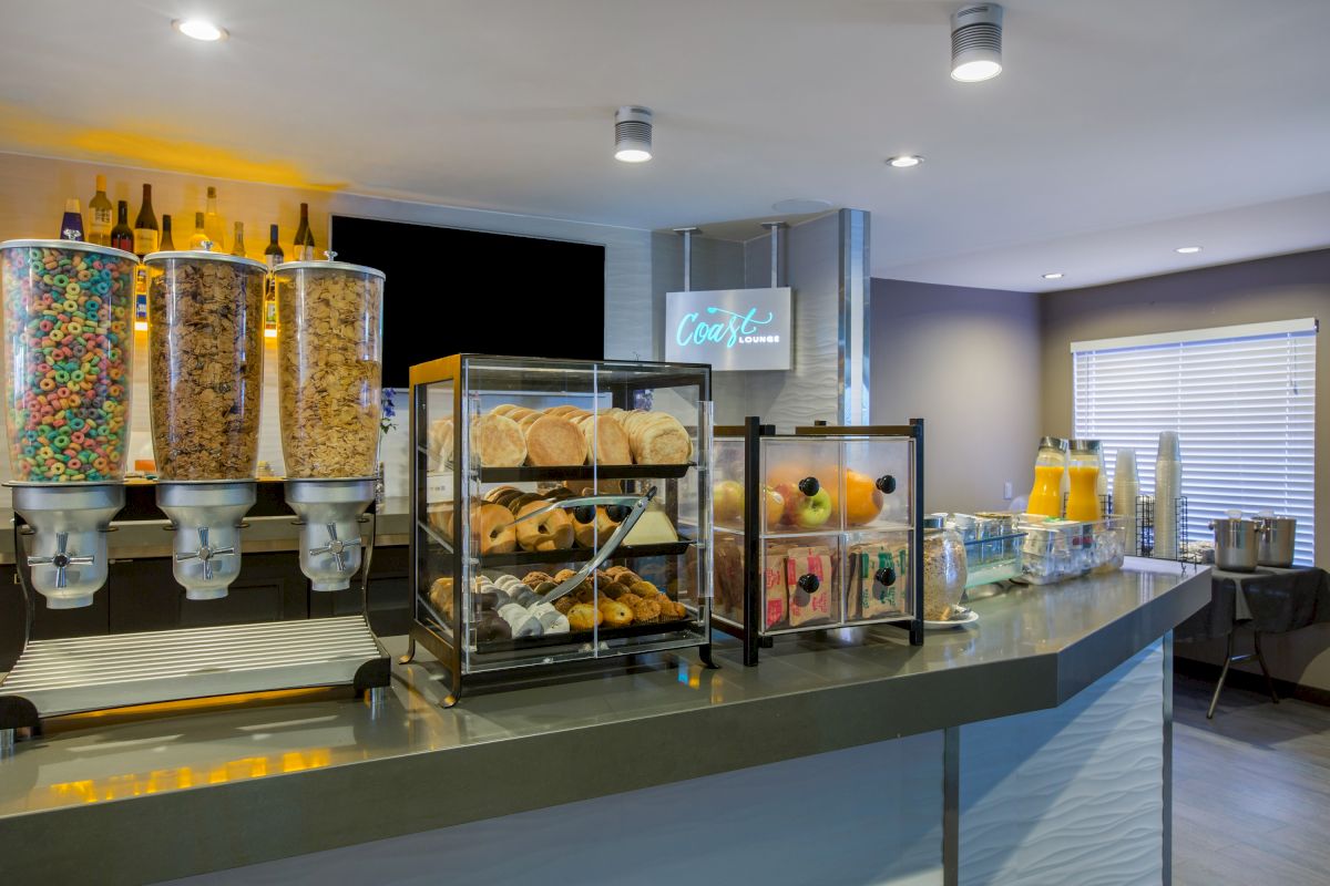 A breakfast buffet at OC Hotel Costa Mesa features cereal dispensers, pastries, bagels, fresh fruit, juice dispensers, and a variety of breakfast items displayed on a counter in a brightly lit room.