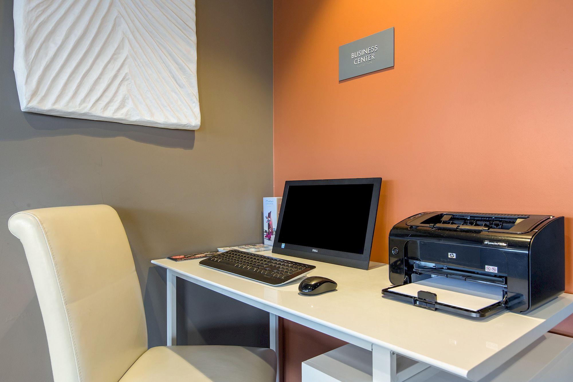 A small business center at OC Hotel Costa Mesa features a white desk, beige chair, computer monitor, keyboard, mouse, black printer, and brochure holder, set against orange and gray walls with a Business Center sign.