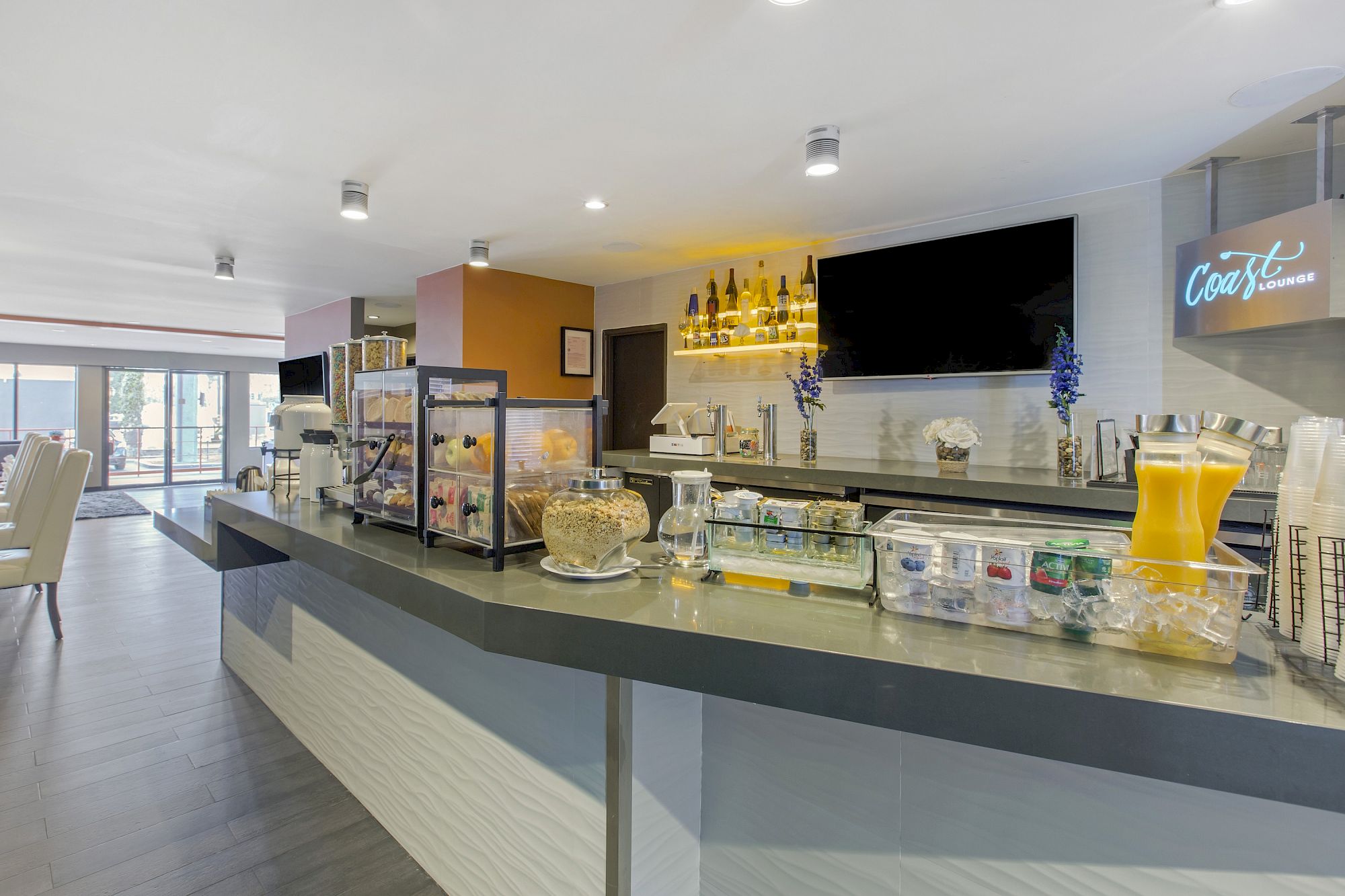 Modern OC Hotel Costa Mesa breakfast bar with cereal dispensers, juice, coffee, and refrigerated drinks on a sleek counter; seating area and large windows in the background. Bottles and a TV are mounted on the back wall.