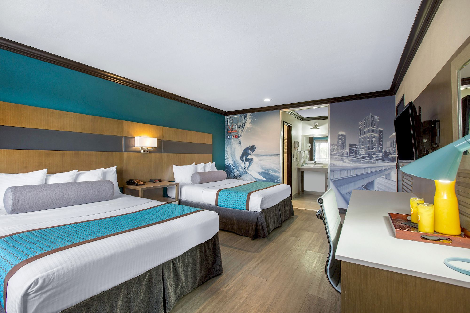 Modern OC Hotel Costa Mesa room with two beds, teal and wood accents, surf-themed wall art, a desk with lamp and yellow drinks, flat-screen TV, and an open bathroom area with cityscape decor.