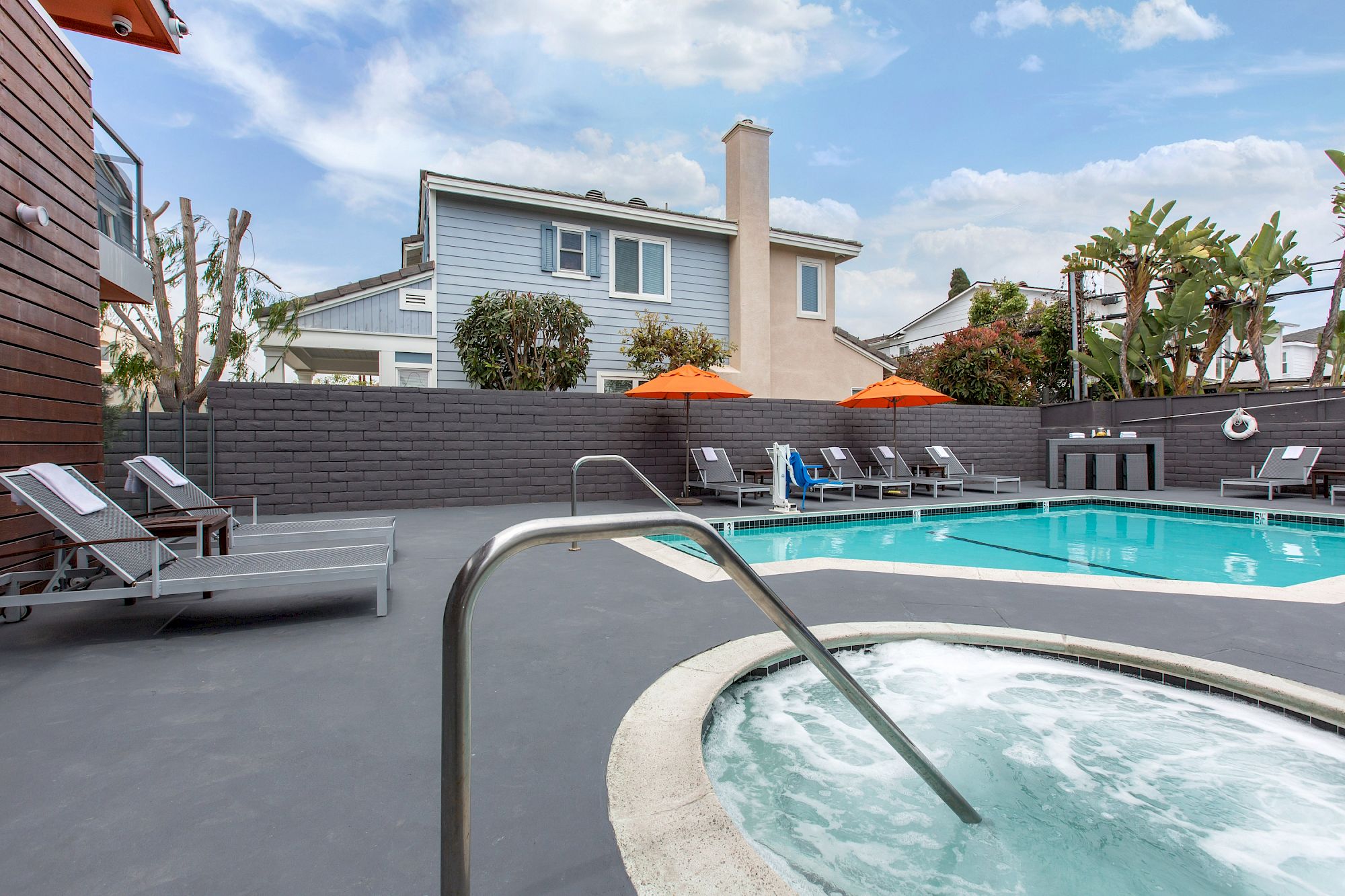 A modern outdoor pool area at OC Hotel Costa Mesa features a hot tub in the foreground, lounge chairs, and orange umbrellas beside a rectangular swimming pool. Residential houses and lush plants are visible under a partly cloudy sky.