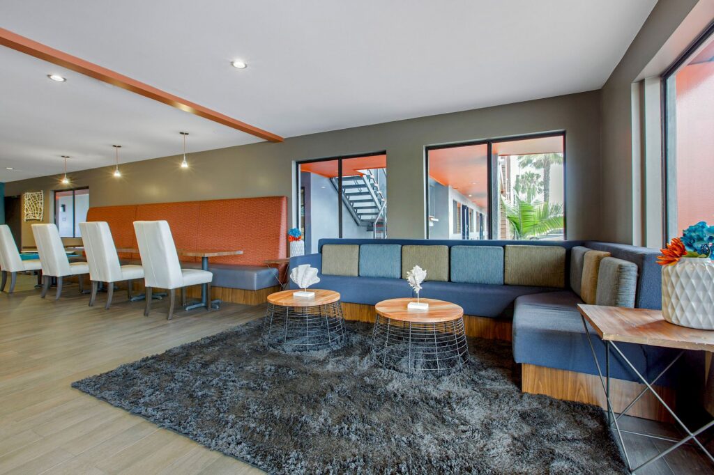 Modern lounge area at OC Hotel Costa Mesa featuring a large L-shaped sofa, two round wooden tables, white chairs at the dining table, and expansive windows that fill the space with natural light. A gray rug adds comfort to the floor.