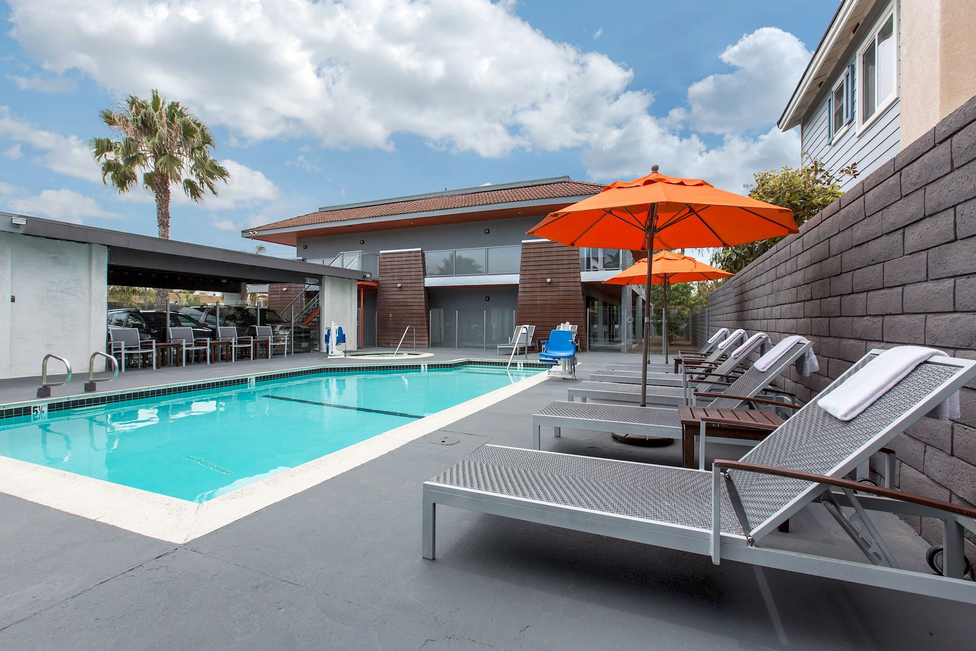 Modern outdoor swimming pool at OC Hotel Costa Mesa with metal lounge chairs under bright orange umbrellas, towels neatly rolled on each chair, and a contemporary building in the background under a partly cloudy sky.