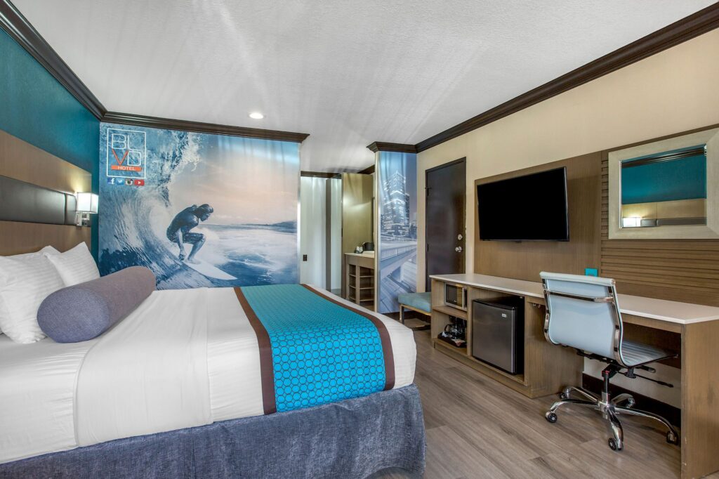 Modern hotel room at OC Hotel Costa Mesa with a bed, blue accents, a large surf-themed mural, flat-screen TV, desk with chair, mini-fridge, and wood flooring. The decor features beach and ocean motifs.