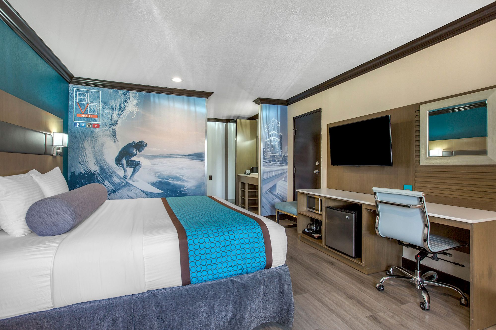 Modern hotel room at OC Hotel Costa Mesa with a bed, blue accents, a large surf-themed mural, flat-screen TV, desk with chair, mini-fridge, and wood flooring. The decor features beach and ocean motifs.