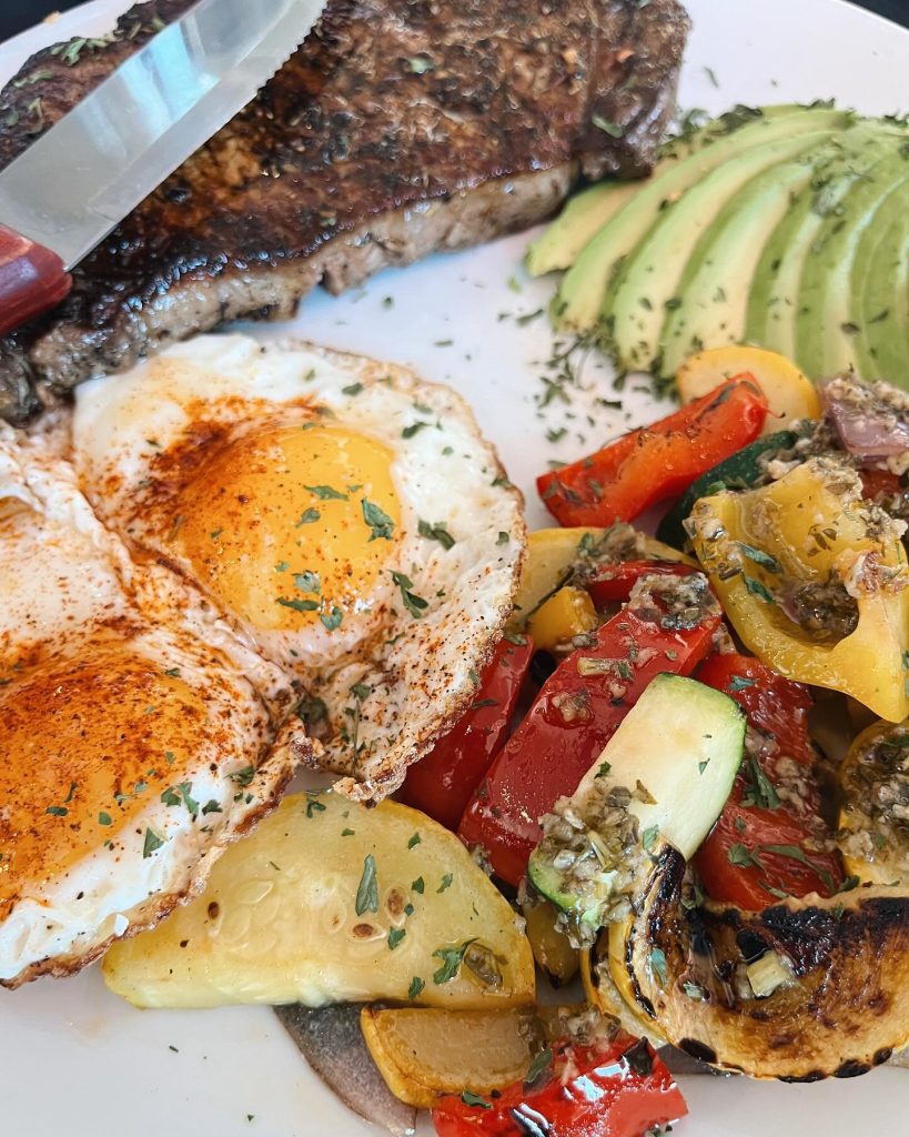 A plate with two sunny-side-up eggs, grilled steak, sliced avocado, and a colorful mix of sautéed vegetables including zucchini and red peppers, garnished with herbs.