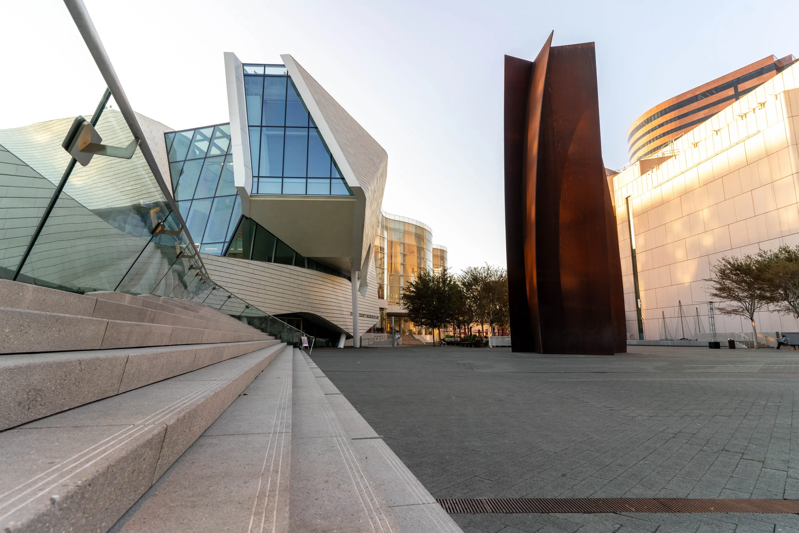 Modern architectural building with glass and angular designs beside wide stone steps, an abstract tall connector sculpture, and trees in a spacious urban plaza under a clear sky in Costa Mesa.