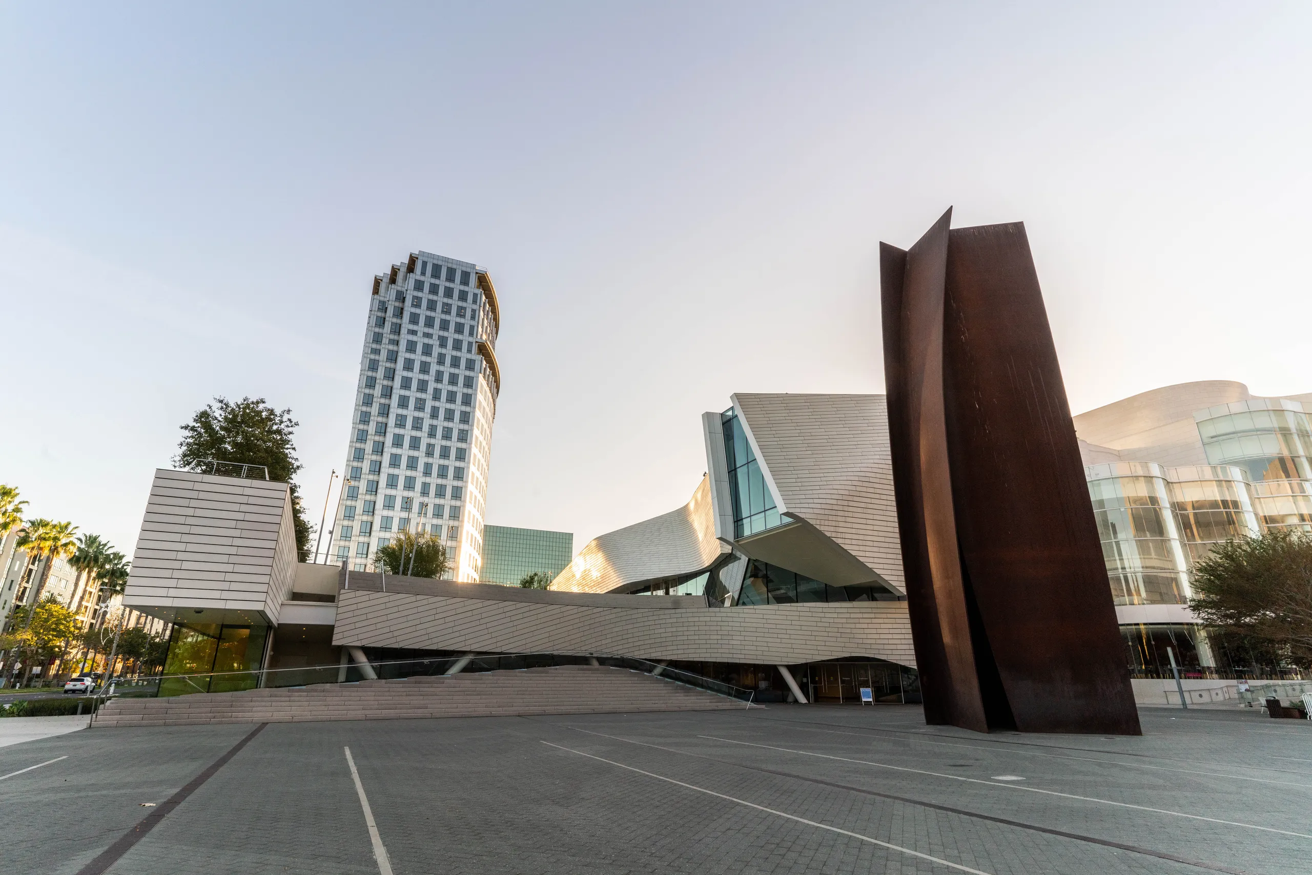 Modern city plaza with the iconic Connector Sculpture of Costa Mesa in the foreground, a unique angular building behind it, and a tall glass skyscraper to the left, all under a clear sky.