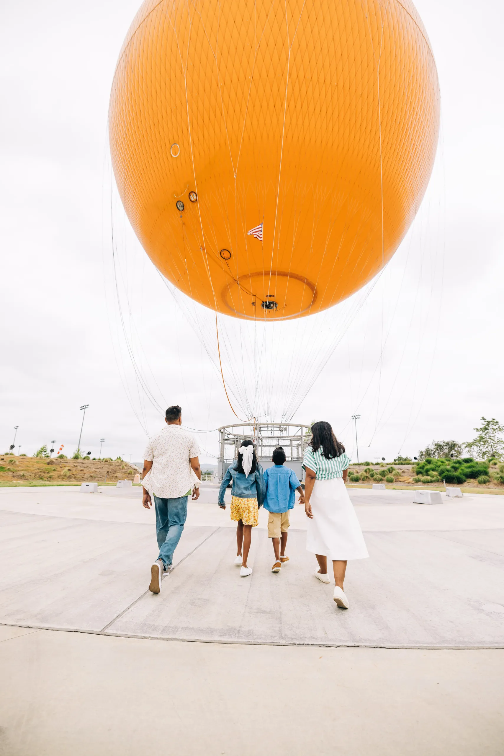 A family of four, seen from behind, walks toward a large orange hot air balloon on a cloudy day in the open park area of Orange County Great Park.