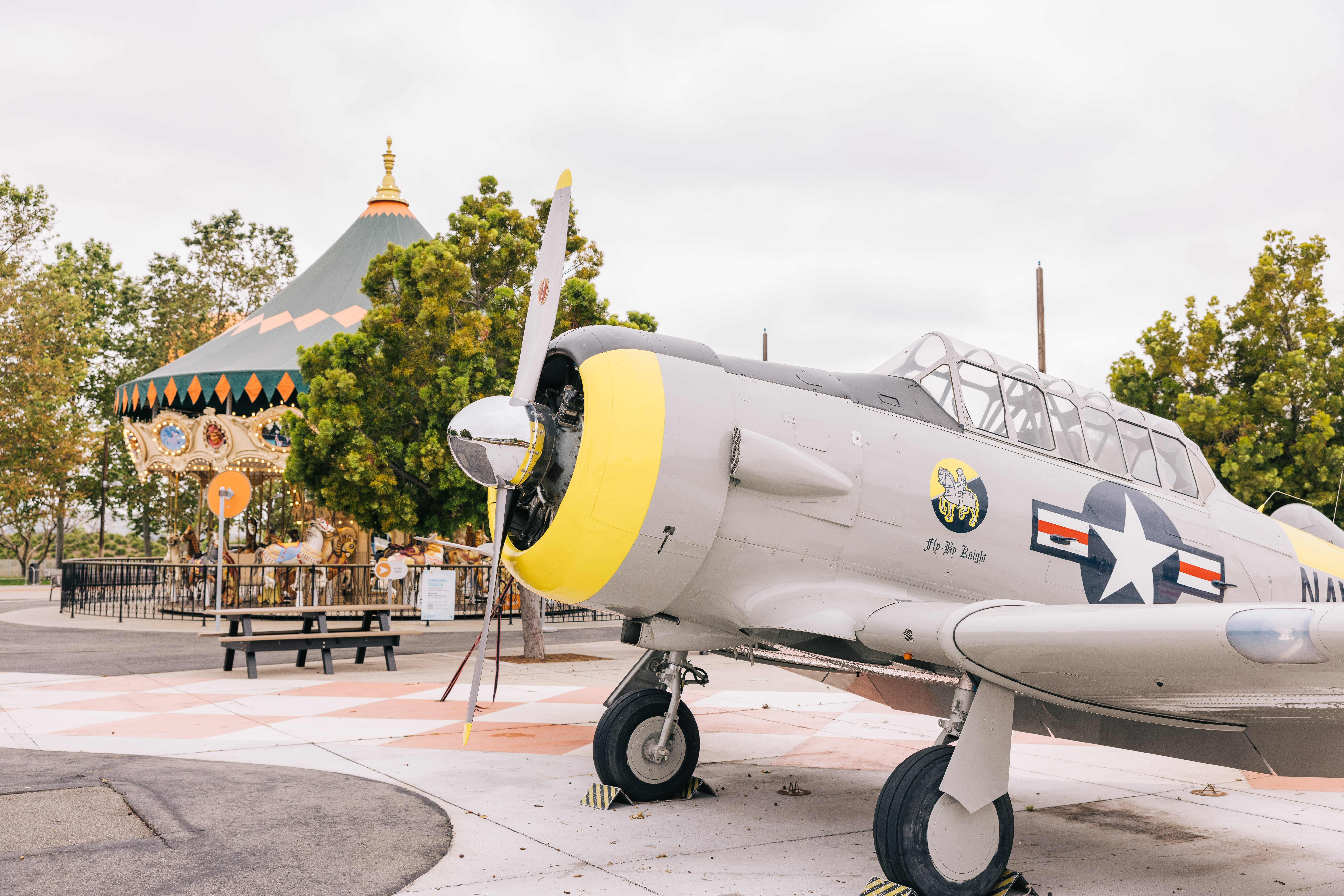 A vintage military aircraft with a yellow nose is displayed outdoors at Orange County Great Park, near a colorful carousel, surrounded by trees and picnic tables under a cloudy sky.