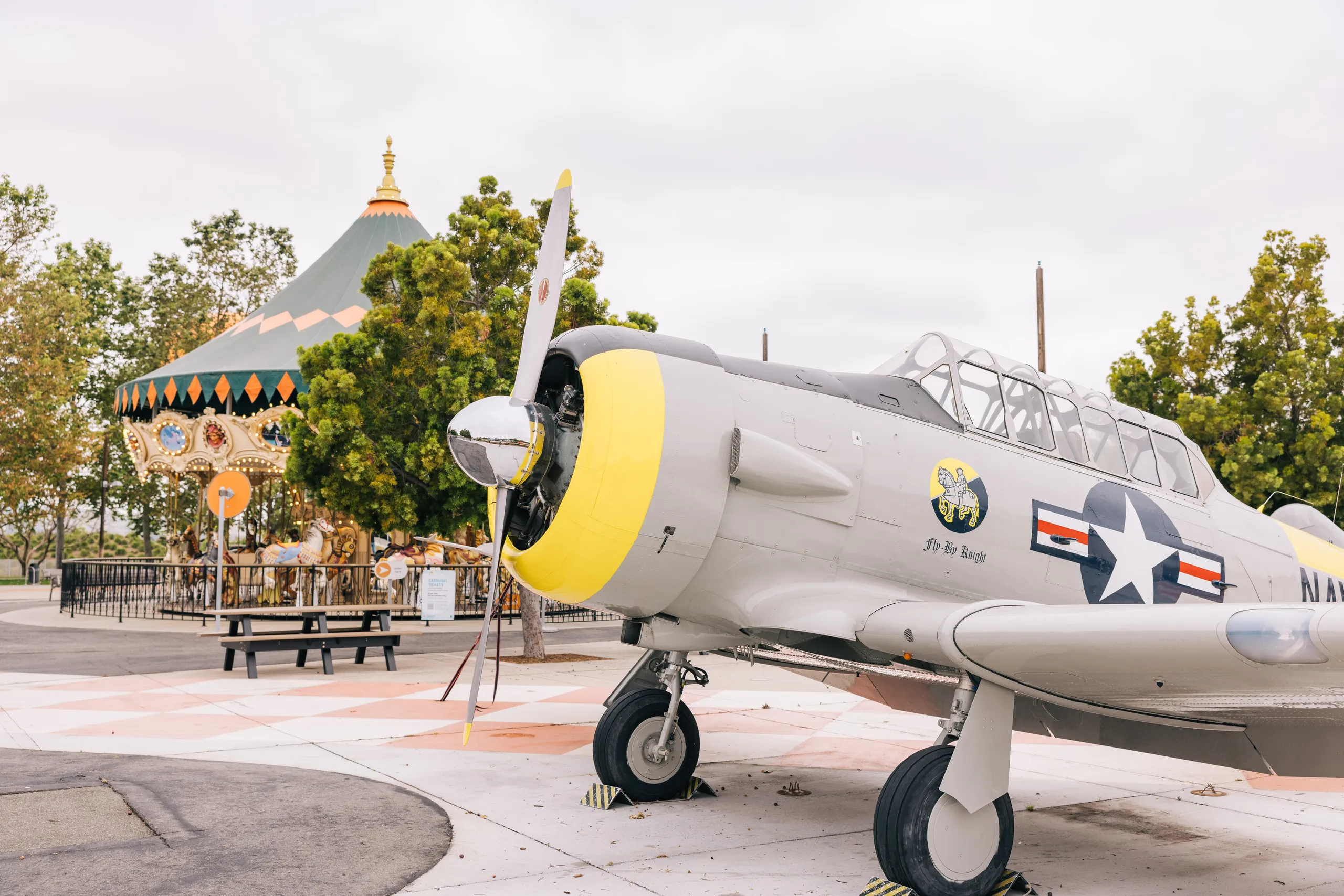 A vintage military aircraft with a yellow nose is displayed outdoors at Orange County Great Park, near a colorful carousel, surrounded by trees and picnic tables under a cloudy sky.