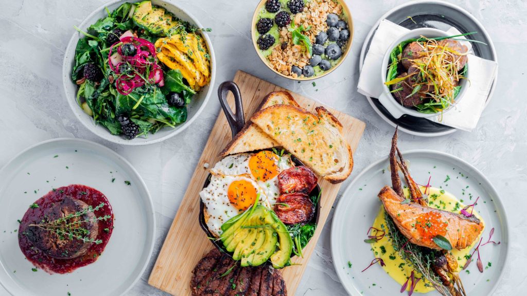 A colorful assortment of dishes including salad bowls, steak with sauce, grilled chicken, eggs with toast, avocado, vegetables, and a bowl of granola with berries, all artfully arranged on a white tabletop.