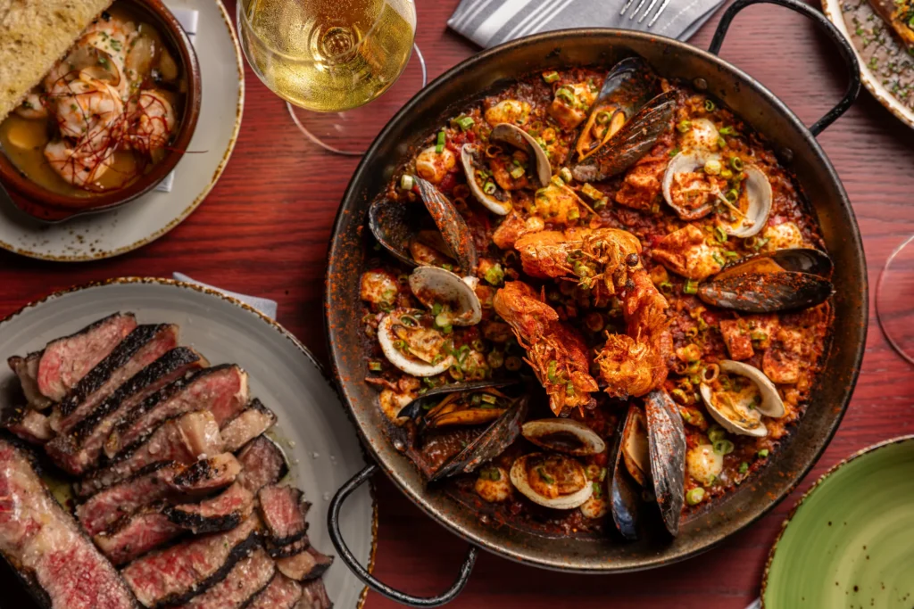 A large pan of seafood paella with shrimp, mussels, and clams sits on a wooden table at Vaca Restaurant Costa Mesa, surrounded by sliced steak, bread, and a glass of white wine.