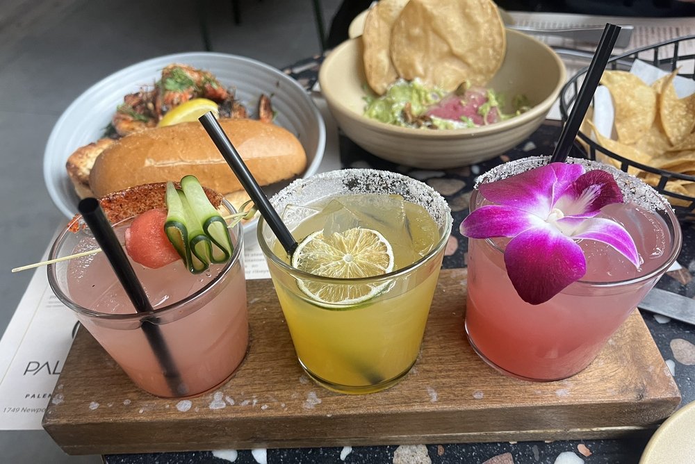 Three colorful cocktails garnished with cucumber, lime, and a purple orchid sit on a wooden tray, with plates of food including chips, guacamole, and a sandwich in the background.