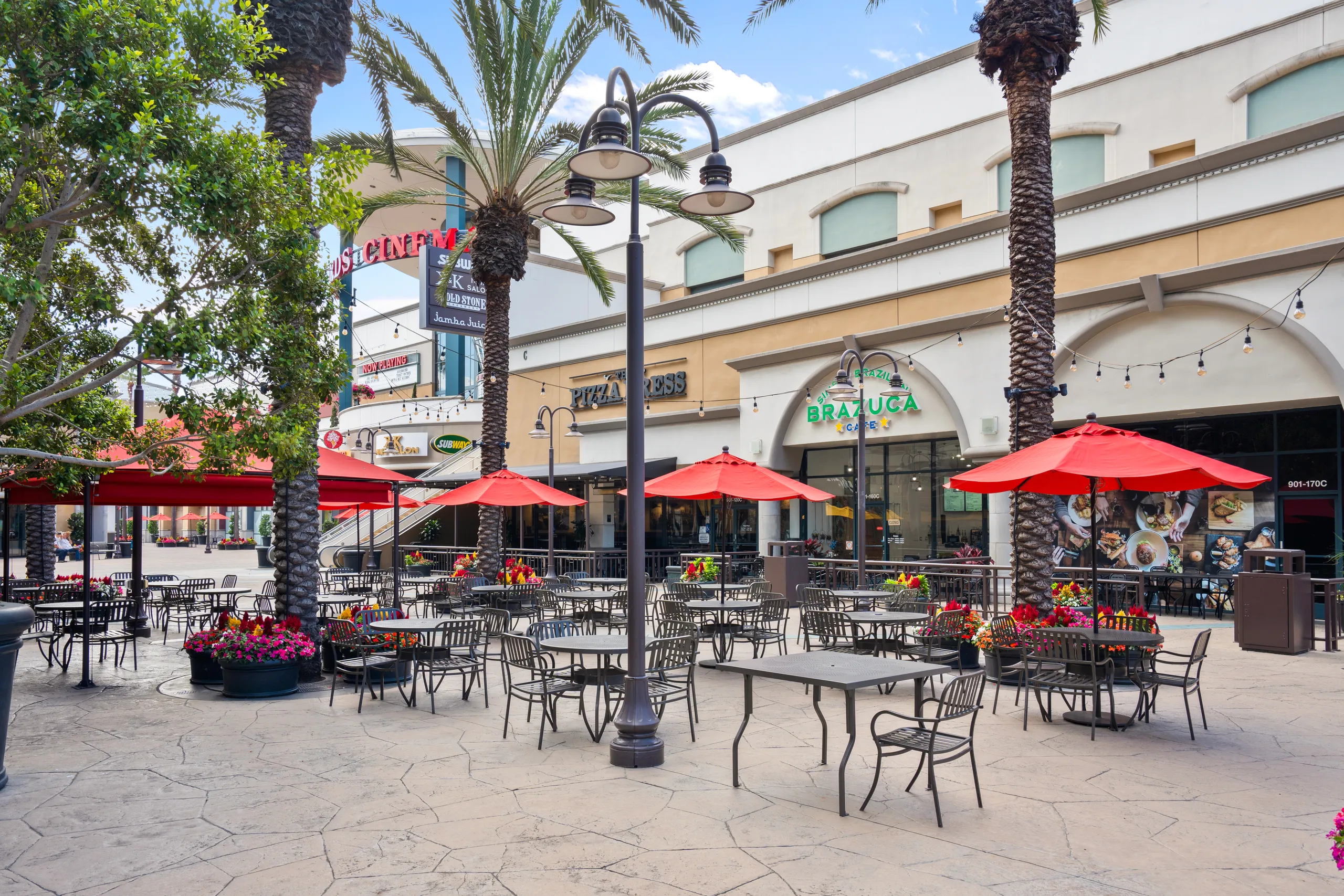 Outdoor patio with empty tables and red umbrellas, surrounded by palm trees and colorful flower pots, in front of shops and restaurants at Metro Pointe Costa Mesa on a sunny day.
