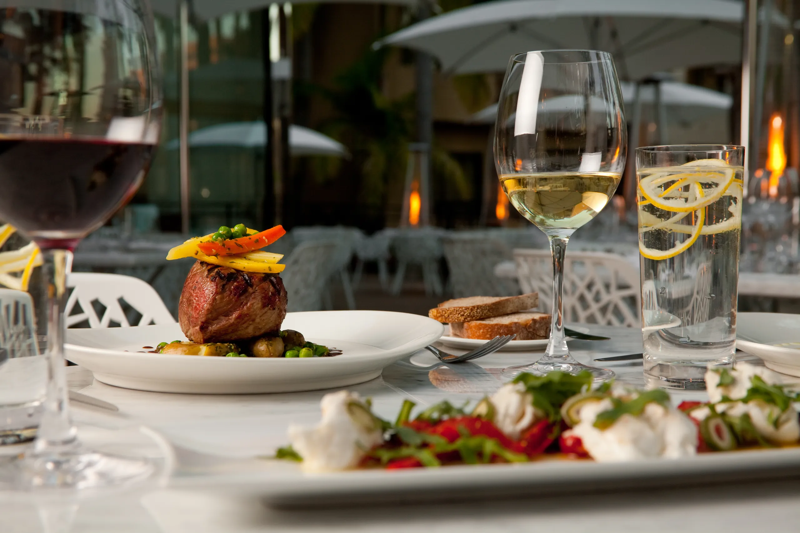 A table set outdoors at Silver Trumpet Restaurant Costa Mesa features gourmet dishes, a glass of red wine, a glass of white wine, and water with lemon slices. White chairs, tables, and umbrellas complete the inviting scene.