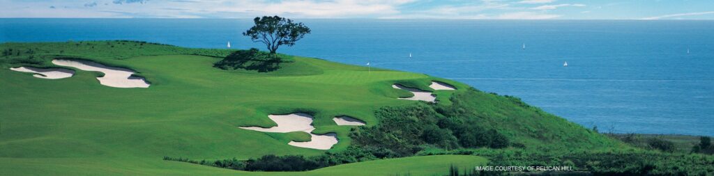 A lush green golf course with sand bunkers overlooks a blue ocean under a partly cloudy sky. A lone tree stands near the top of a hill, and small sailboats are visible on the water in the distance.