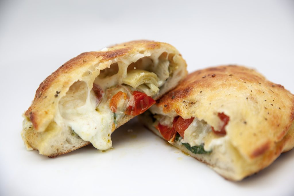 A golden-brown calzone cut in half, revealing melted cheese, artichoke hearts, tomatoes, basil, and other vegetables inside, all on a plain white background.
