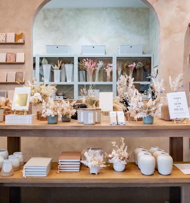 A neatly arranged shop display features white vases, dried floral bouquets, notebooks, and greeting cards on wooden shelves, with shelves of more vases and flowers in the background under a wide archway.