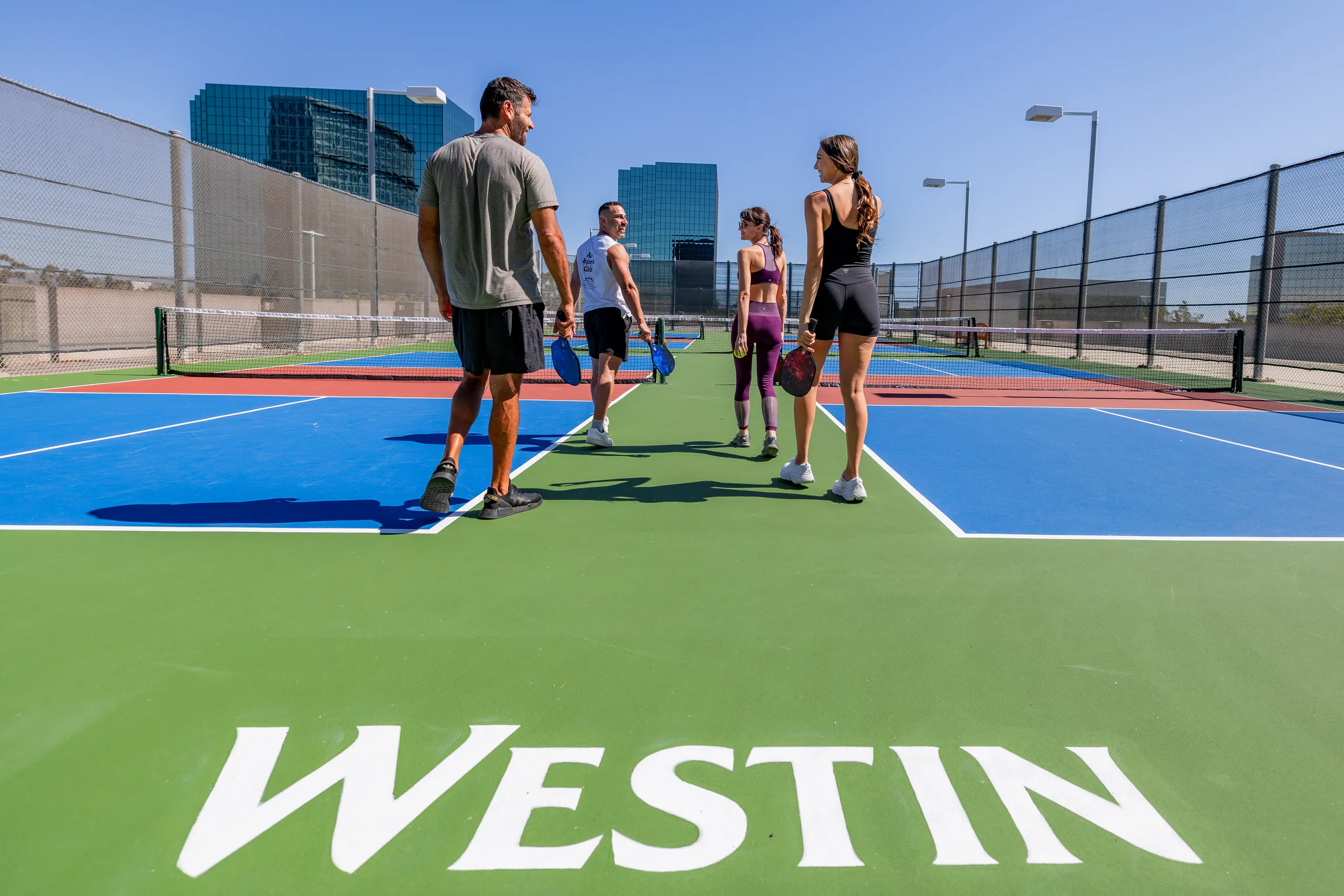 Four people with pickleball paddles walk on an outdoor court with “WESTIN” written on the ground at Westin South Coast Plaza. Tall buildings and a clear blue sky are visible in the background.