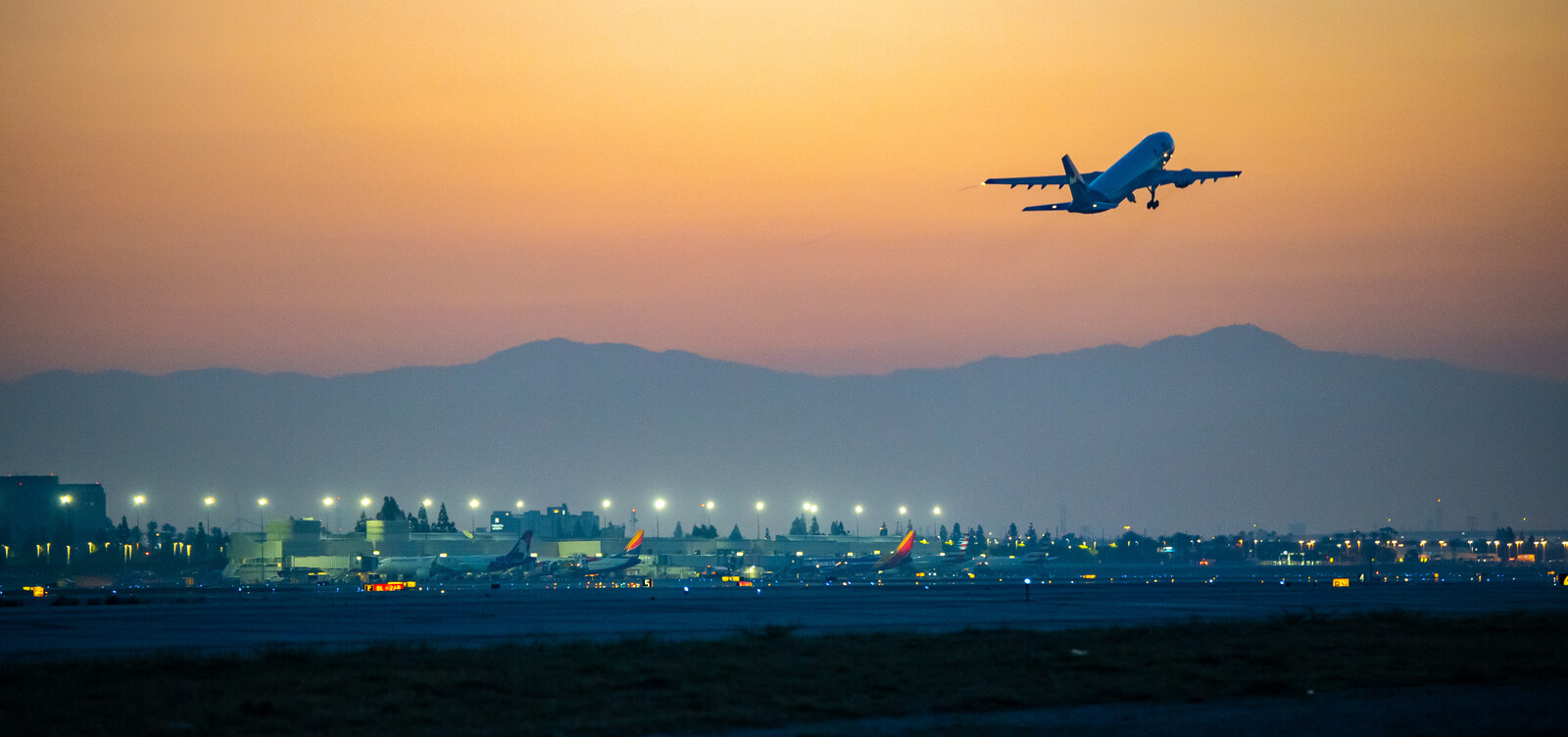 An airplane takes off at sunset from a brightly lit airport, with terminal buildings, parked planes, and mountains in the background—perfect for travelers getting to Costa Mesa from the airport under an orange and blue sky.