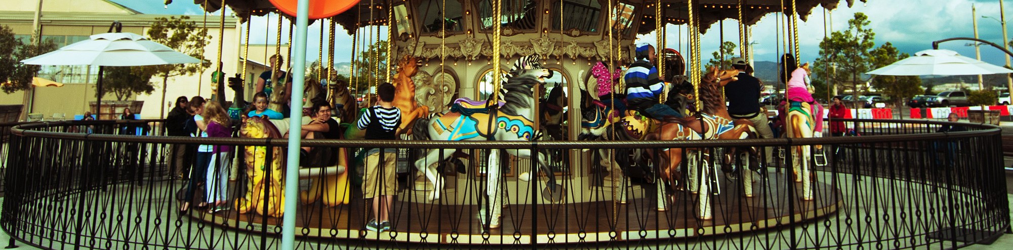 Children and adults ride colorful horses on a merry-go-round carousel at Orange County Great Park, with onlookers standing nearby behind a black fence. White umbrellas and trees are visible in the background.