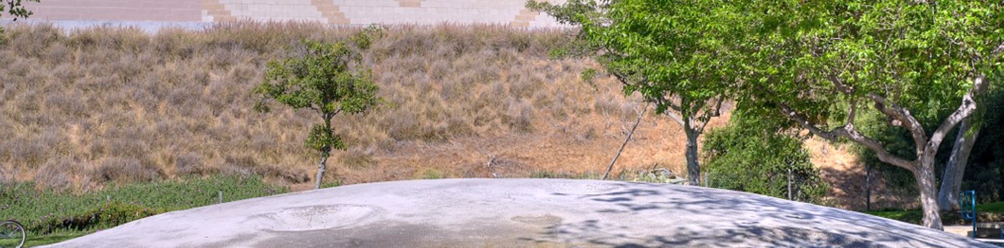 A large, smooth, dome-shaped rock at Moon Park Costa Mesa is surrounded by trees and dry grass, with a beige wall in the background and sunlight casting shadows on the scene.