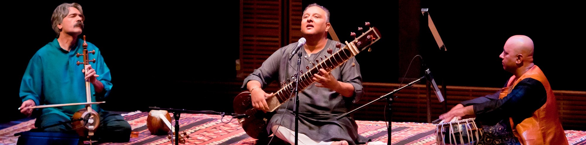 Three musicians sit cross-legged on a patterned rug, performing on stage for the Philharmonic Society Orange County. One plays a bowed string instrument, another the sitar, and the third tabla drums, all deeply focused and immersed in the music.