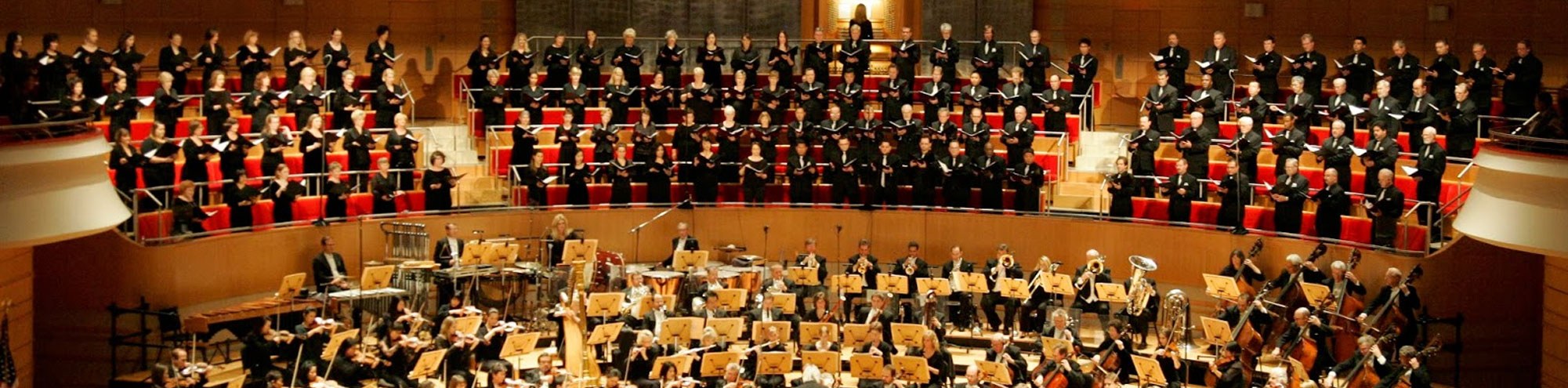The Pacific Symphony Orange County performs on stage with a full choir standing in tiered rows. Musicians play various instruments, and the choir members hold sheet music in a brightly lit concert hall.