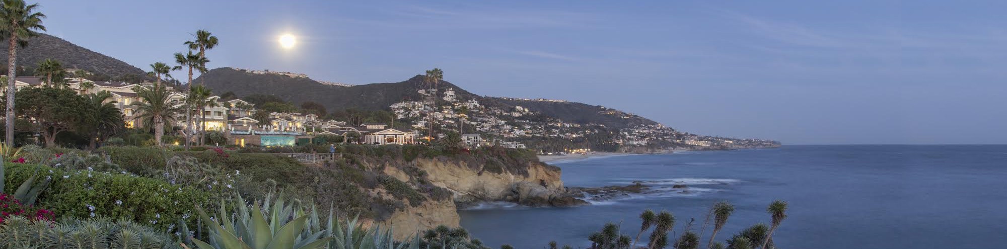 A coastal scene at dusk in Laguna Beach, California shows houses on cliffs overlooking the ocean, with palm trees and lush greenery in the foreground and a bright, full moon rising over distant hills.