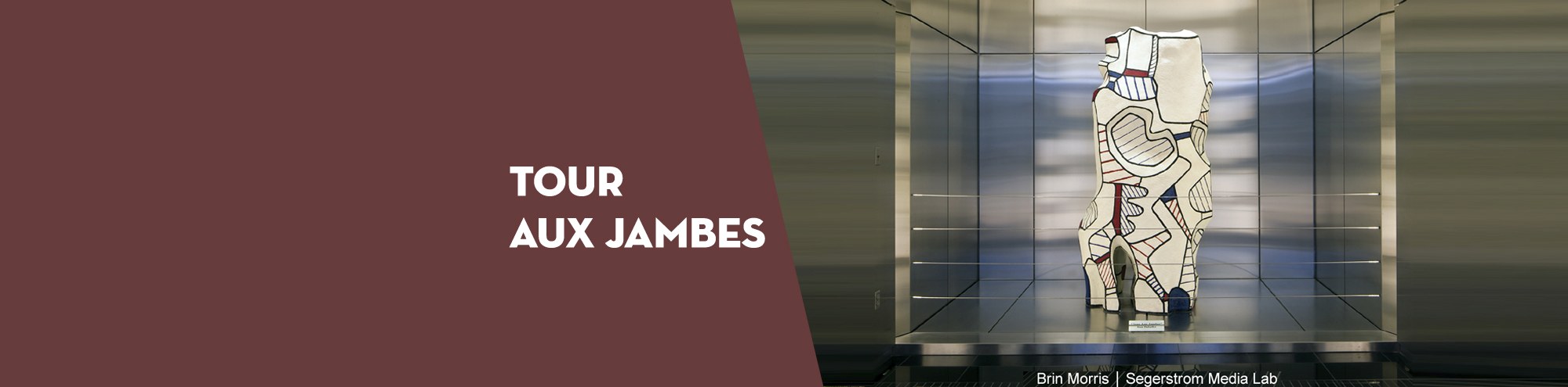A modern abstract sculpture with bold lines and shapes is displayed in a glass case. Beside it, text on a maroon background reads TOUR AUX JAMBES, highlighting this Costa Mesa installation by Sir Morris | Spacestorm Media Lab.