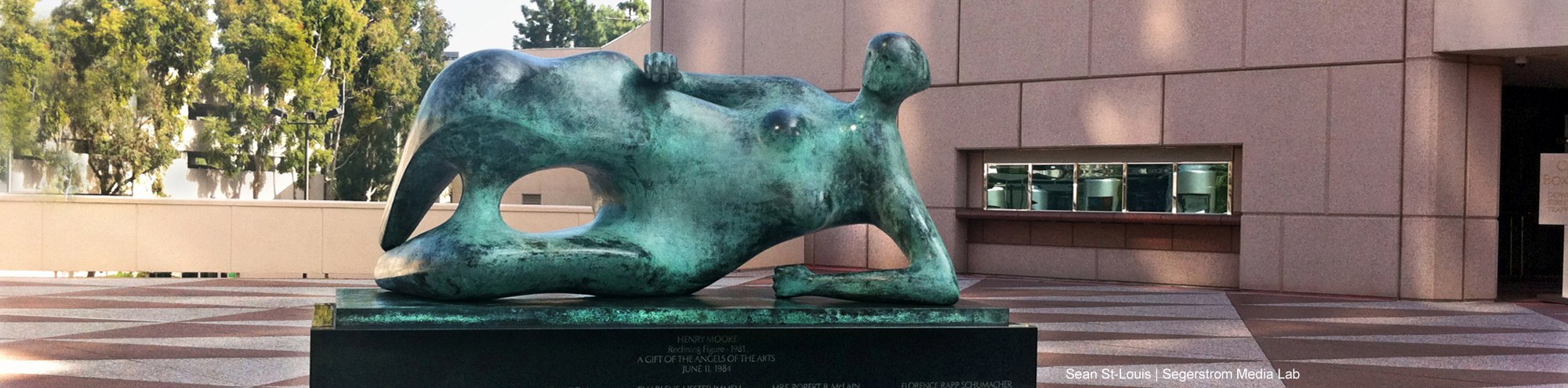 A large bronze reclining figure sculpture graces a geometric-patterned plaza in Costa Mesa, set against modern buildings and leafy trees.