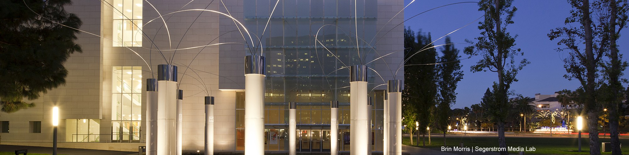 Modern building at dusk with large glass windows, tall white columns, and the utsurohi 91 costa mesa metal sculptures arching overhead. Trees and streetlights line the surrounding area.