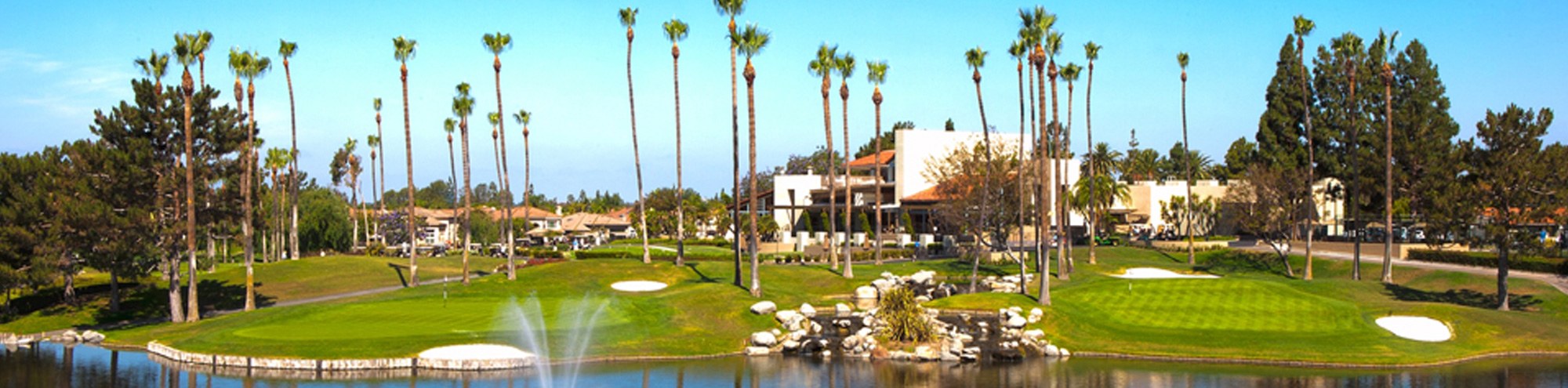 A golf course with lush green lawns, sand traps, tall palm trees, a small pond with a fountain, and a clubhouse in the background under a clear blue sky.