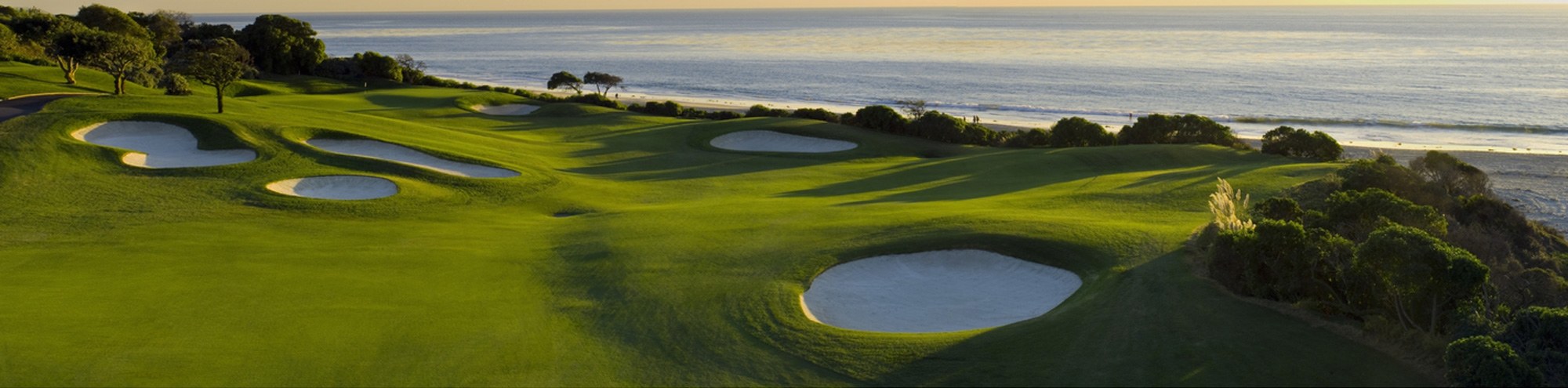 A scenic golf course with lush green fairways, several sand bunkers, and trees, set along a coastline with the ocean and gentle waves visible in the background at sunset.