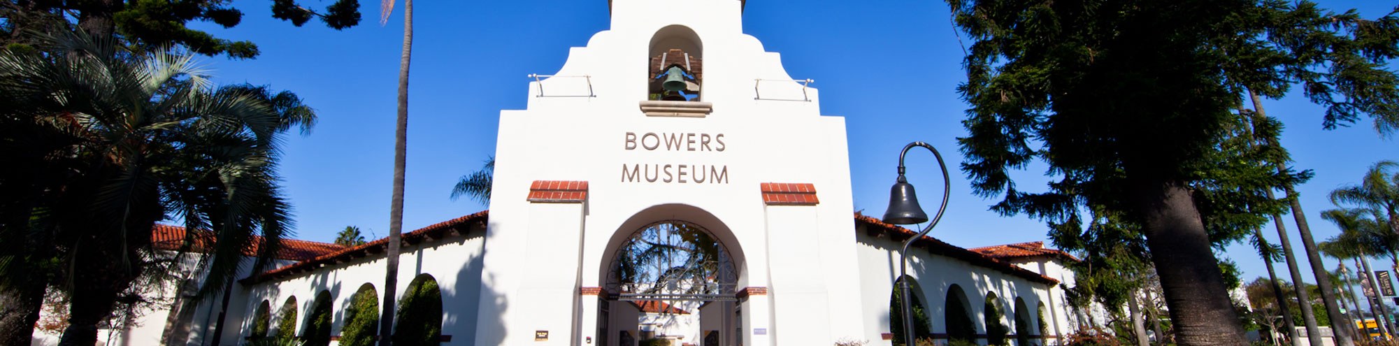 The Bowers Museum Orange County’s white stucco entrance features an arched doorway, a bell tower, and the museum’s name above the arch, surrounded by tall palm trees and greenery under a clear blue sky.