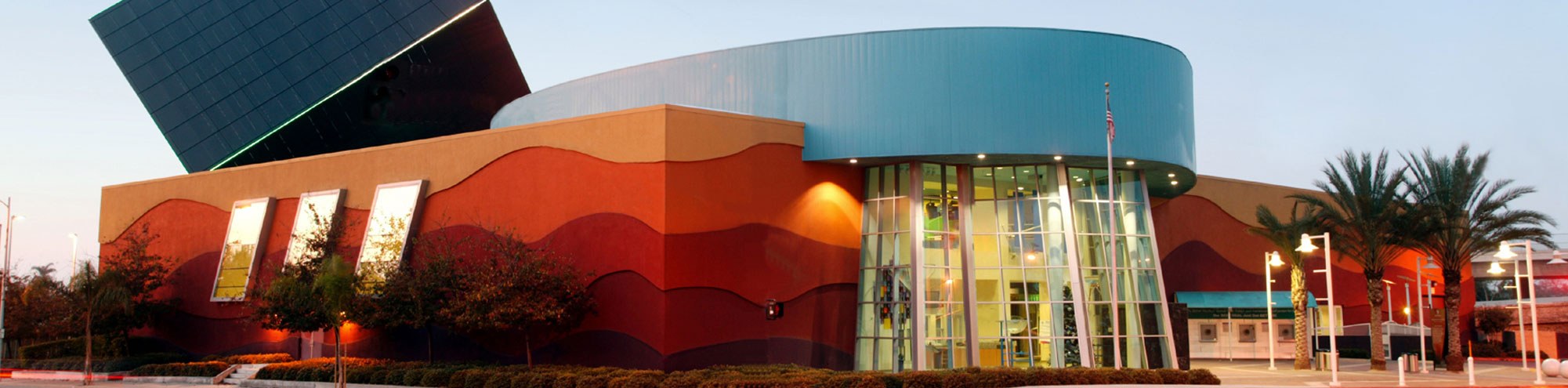A modern building with bold, colorful wave patterns on its exterior walls, tall glass entrance, and geometric shapes, surrounded by palm trees and landscaping under a clear sky.