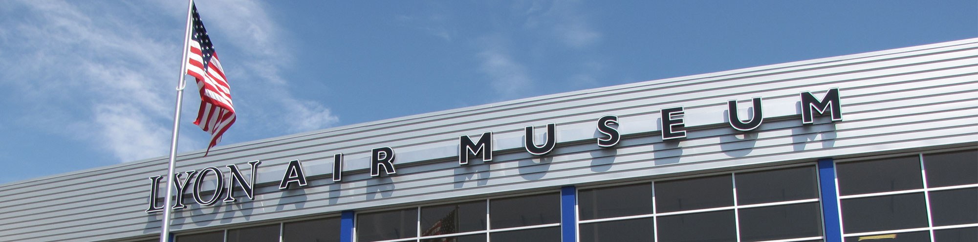 Exterior view of Lyon Air Museum with large black letters on a silver building facade and an American flag on a tall flagpole against a blue sky with wispy clouds.
