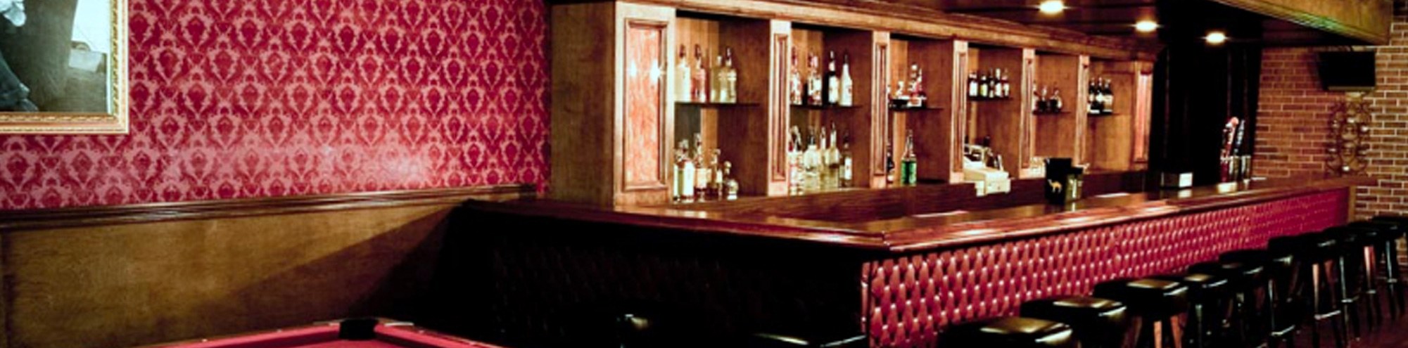 A dimly lit bar with red patterned wallpaper, a wooden counter with red tufted panels, rows of bar stools, shelves of liquor bottles behind the bar, and part of a pool table in the foreground.