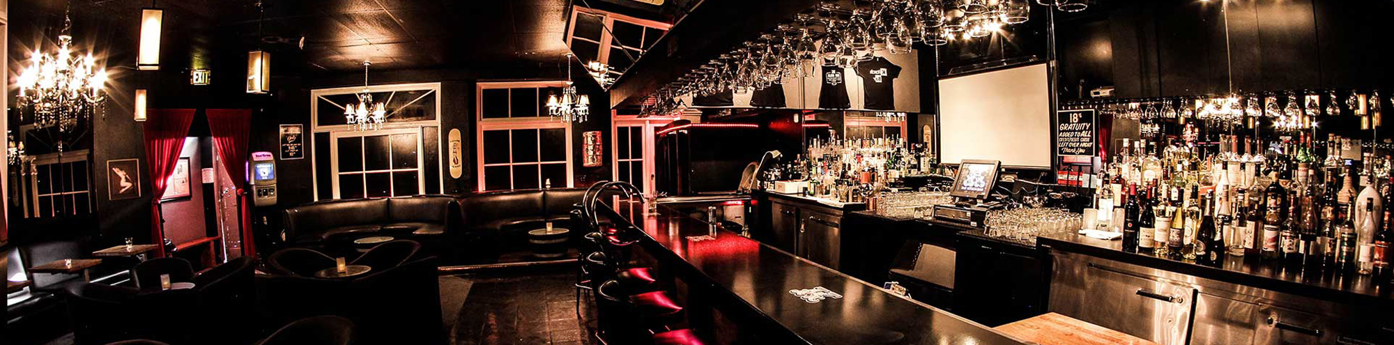 A dimly lit, stylish kitsch bar in Costa Mesa with black leather seating, chandeliers, a long counter lined with stools, and shelves full of liquor bottles behind the bar. Glasses hang upside down above the counter.