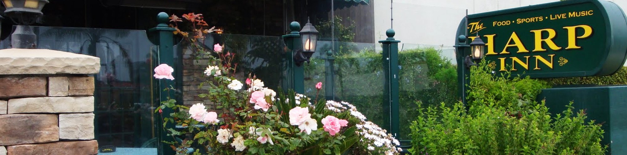Outdoor view of The Harp Inn pub sign with flowers and greenery in the foreground, glass panels, and lantern-style lights creating a welcoming entrance.