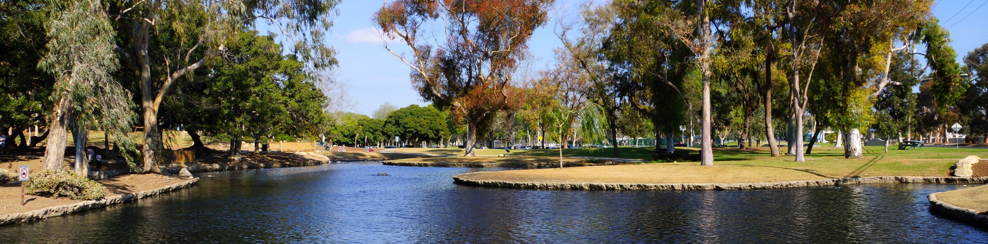 A peaceful park scene at TeWinkle Park, Costa Mesa, with a winding river, grassy banks, and tall trees under a clear blue sky. People can be seen walking and relaxing in the background.