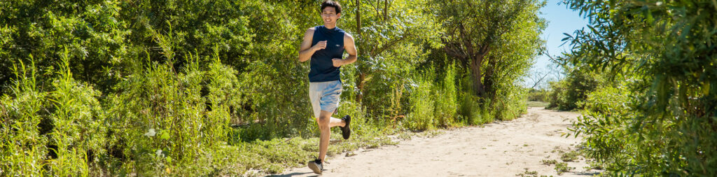 A young man jogs along a sandy path surrounded by lush green trees and shrubs under a clear blue sky at Talbert Nature Preserve in Costa Mesa.