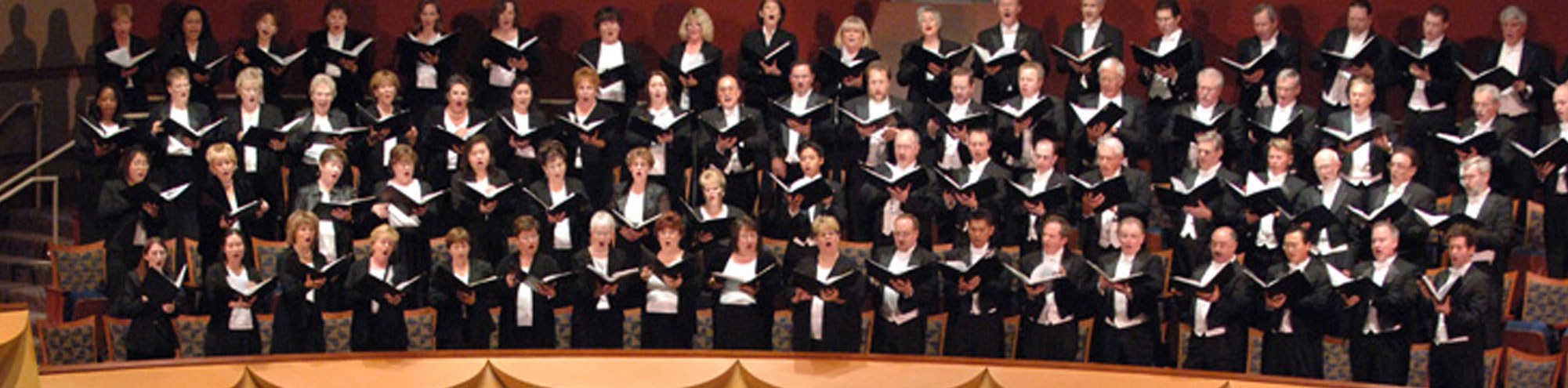 The Pacific Chorale Orange County, dressed in black and white robes, stands on tiered risers in a concert hall, holding open music folders and singing together.