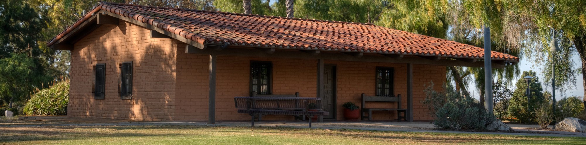 A small, rustic adobe house with a red tile roof sits in a grassy area surrounded by trees. Benches and potted plants grace the shaded porch, as late afternoon sunlight casts soft shadows on the historic Diego Sepulveda Adobe in Costa Mesa.