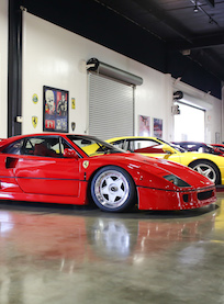 A red Ferrari sports car is parked indoors next to a yellow Ferrari and other cars, with posters on the white walls and a glossy concrete floor reflecting the vehicles.