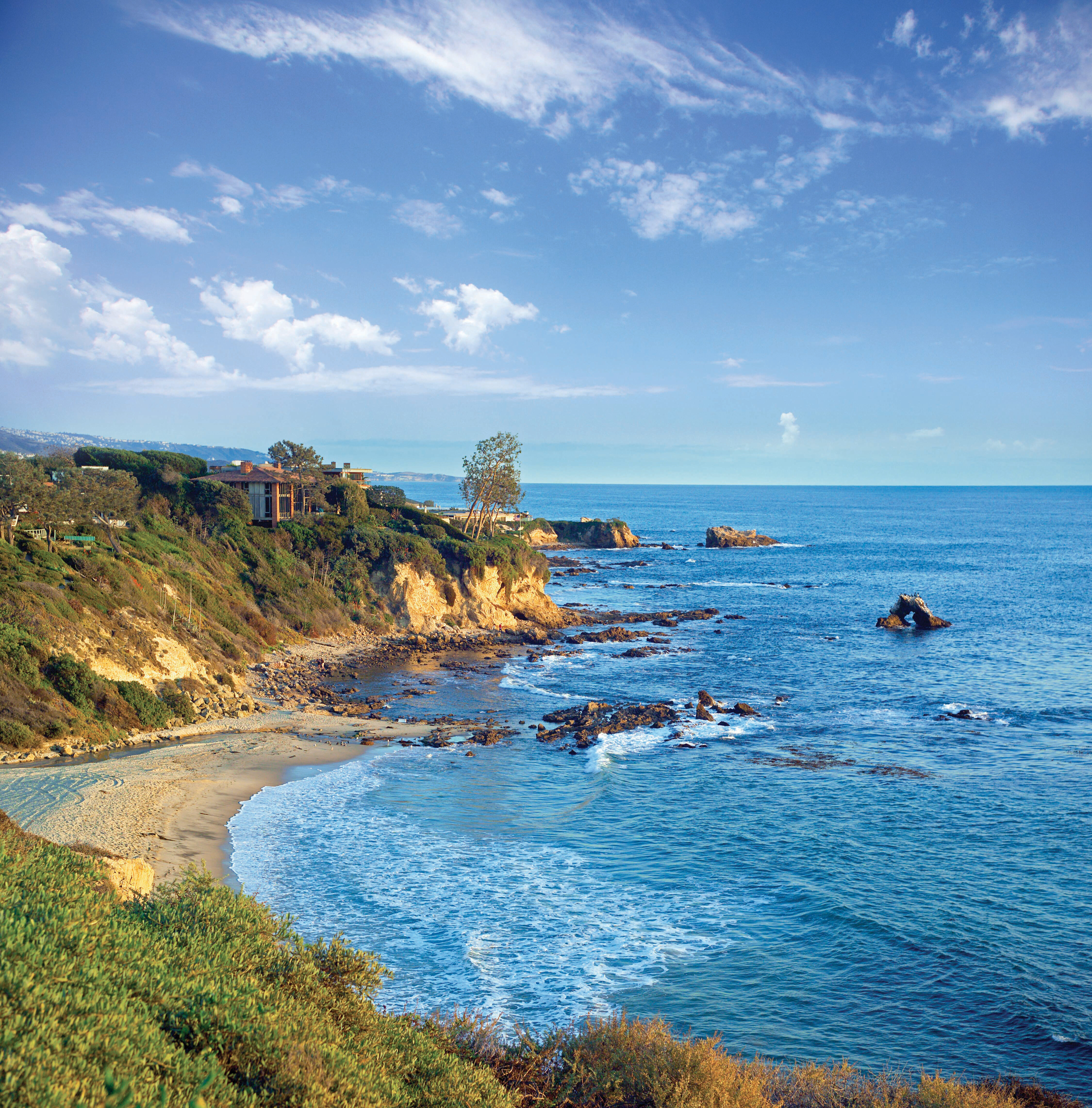 A scenic coastal view with cliffs, a sandy beach, rocky shoreline, and blue ocean waves under a partly cloudy sky. Some houses and greenery are visible atop the cliffs.