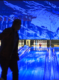 A person is silhouetted in the foreground while bowling in a glowing bowling alley with neon blue lighting and pins set up at the end of the lane.