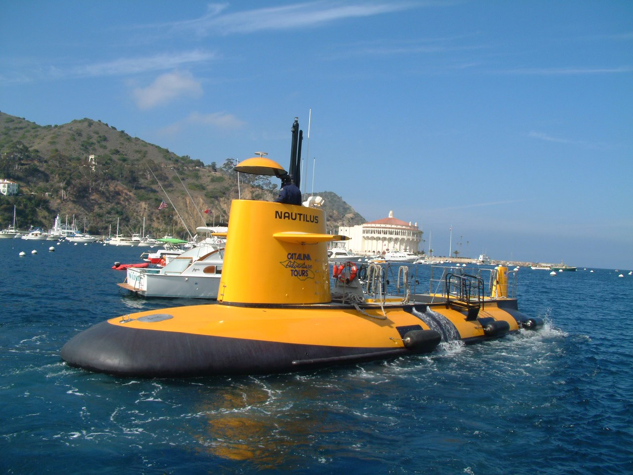 A yellow submarine named Nautilus floats on the blue water near a coastline with boats and a white domed building in the background under a clear sky.