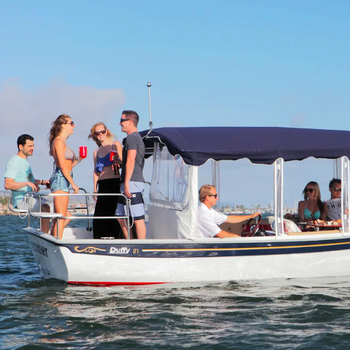 A group of young adults enjoy a sunny day on a small white boat with a navy canopy, talking and relaxing as the boat cruises on calm water. The driver steers while others stand or sit, holding drinks.